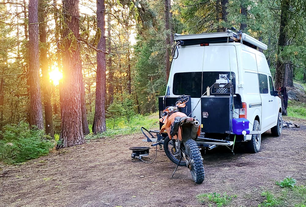 van and motorycle parked in a forest with setting sun behind