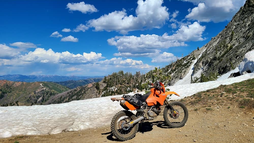 motorcycle parked next to a snow patch with sweeping vista behind