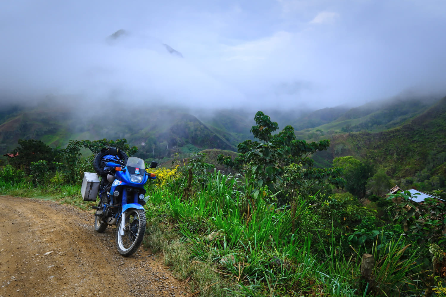 motorcycle parked on a dirt road with low clouds obscuring 
             lush mountains behind