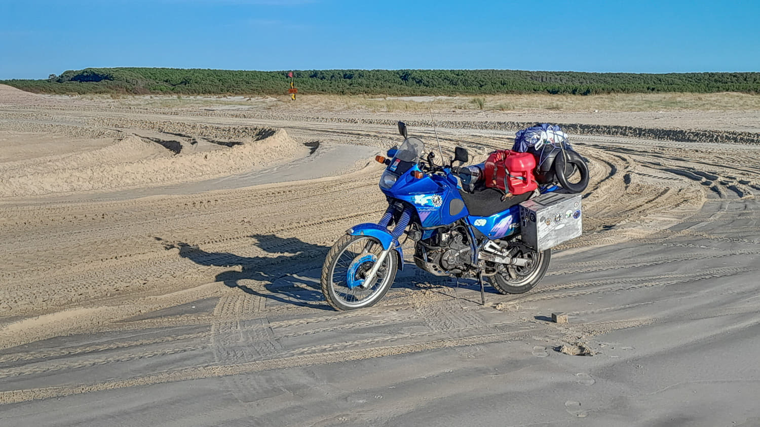 motorcycle overloaded with luggage and a big red gas canister parked on a sandy road