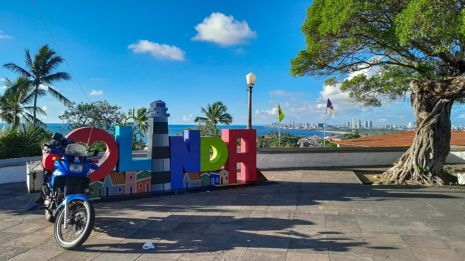 motorcycle parked next to a colorful sign Olinda with blue ocean behind