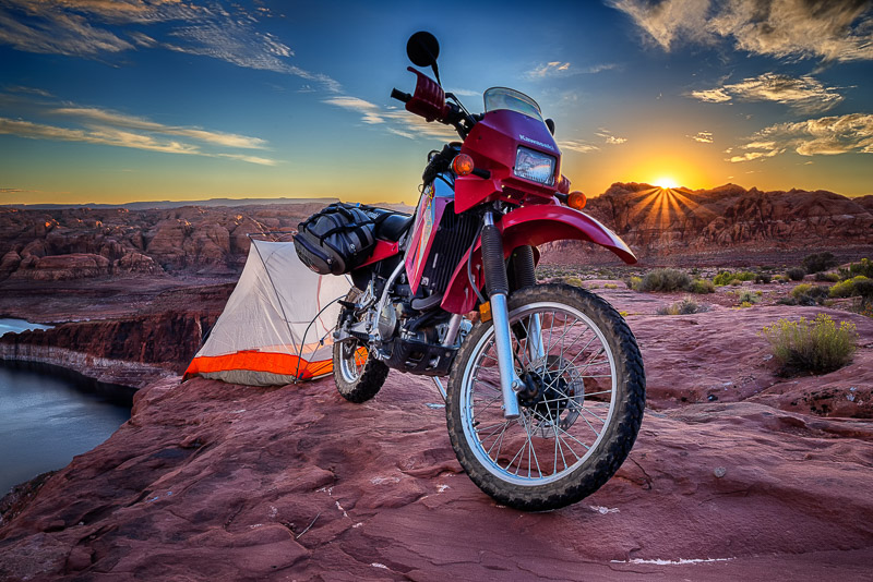 Motorcycle and a tent perched on a cliffside with first rays of raising sun peaking over
			remote peaks