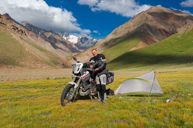 posing next to a tent and motorcycle with snow covered mountains behind