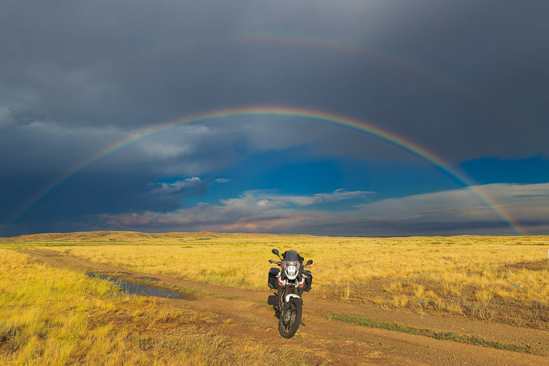 motorcycle parked on a yellow-grass field with rainbow against dark sky behind