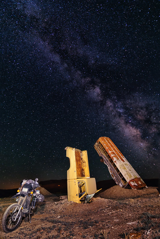 motorcycle in front of a pick-u and schooo bus half-burried vertically in the ground
             with milky way in the background