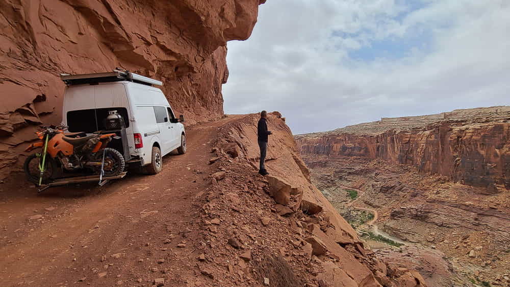 van and person next to it on a narrow dirt road hugging edge of a canyon